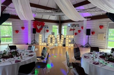 Salle décorée pour un événement avec des tables dressées, des ballons en forme de cœur et le mot ’LOVE’ illuminé.
