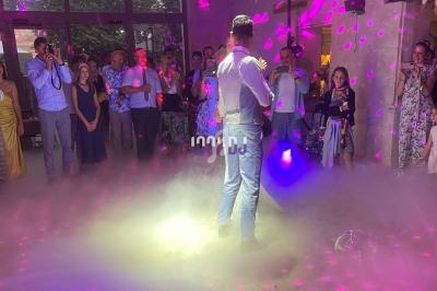 Un homme en costume danse au centre d'une piste entourée d'invités, éclairée par des lumières colorées et de la fumée.