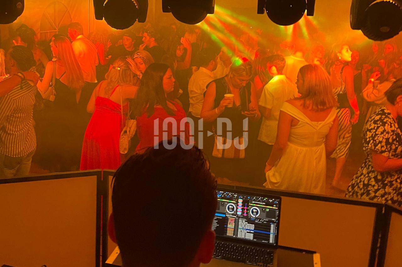 Vue depuis la cabine d’un DJ sur une piste de danse bondée, éclairée par des lumières colorées et une ambiance festive.