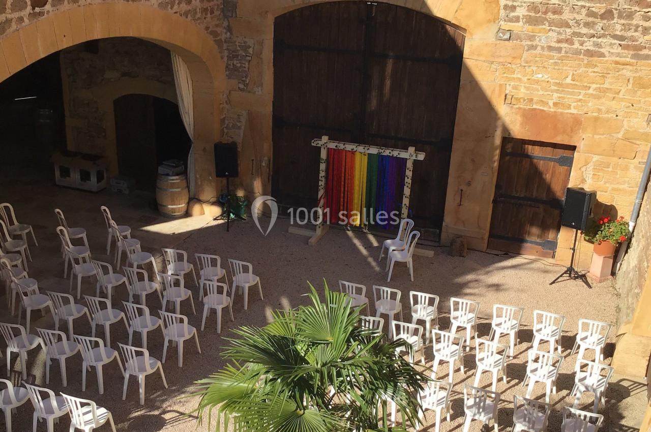 Cour intérieure ensoleillée avec des rangées de chaises blanches disposées face à une scène décorée de rideaux colorés.