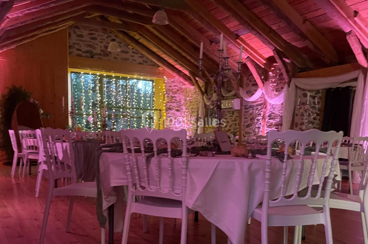 Salle de réception chaleureuse avec tables dressées, chaises blanches et éclairage tamisé aux tons roses.