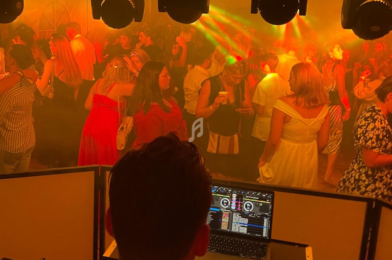 Vue depuis la cabine d’un DJ sur une piste de danse bondée, éclairée par des lumières colorées et une ambiance festive.