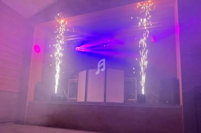 Scène de spectacle avec jeux de lumière violets, étincelles pyrotechniques et fumée dans une salle intérieure.