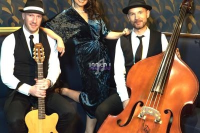 Un trio musical pose avec une guitare, une contrebasse et une chanteuse dans un décor intérieur élégant.