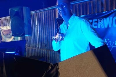 Homme en chemise blanche derrière une table de mixage, éclairé par une lumière bleue, en extérieur la nuit.