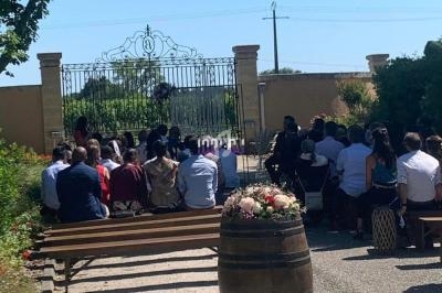 Un groupe de personnes assises en extérieur, face à une grille en fer forgé, lors d'une cérémonie sous un ciel dégagé.