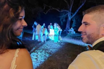 Un couple souriant se regarde lors d'une soirée en extérieur, avec un groupe de personnes en arrière-plan sous des lumières.