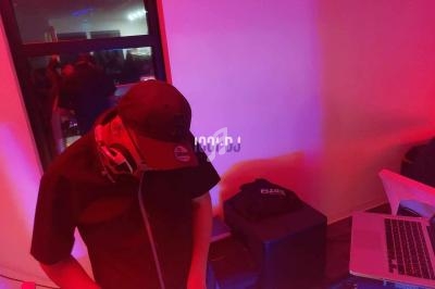 Un DJ portant une casquette mixe de la musique sur une console dans une pièce éclairée par des lumières rouges et violettes.