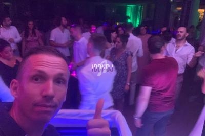 Une foule danse dans une ambiance festive et lumineuse dans un club, avec un homme au premier plan faisant un signe de pouce…