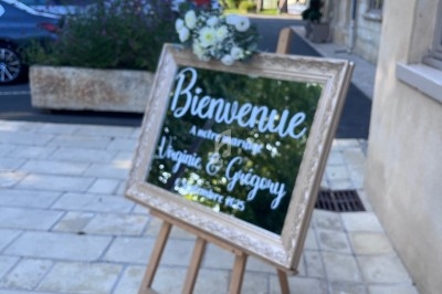 Panneau de bienvenue décoré de fleurs, posé sur un chevalet devant un bâtiment en pierre.