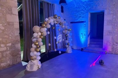Arche décorative en ballons blancs et dorés dans une salle en pierre éclairée par des lumières colorées.