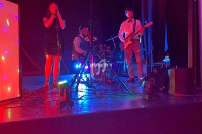 Un groupe de musique joue sur scène avec éclairages colorés, une chanteuse, un batteur et un guitariste visibles.