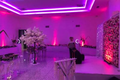 Salle décorée avec des lumières violettes, un arbre artificiel blanc et un mur floral, avec deux personnes présentes.