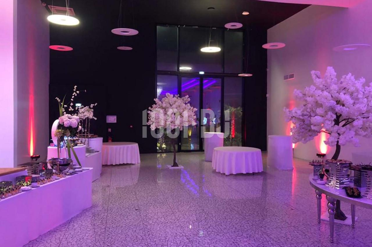Salle de réception décorée avec des tables rondes drapées de blanc, des arbres artificiels et un éclairage coloré.