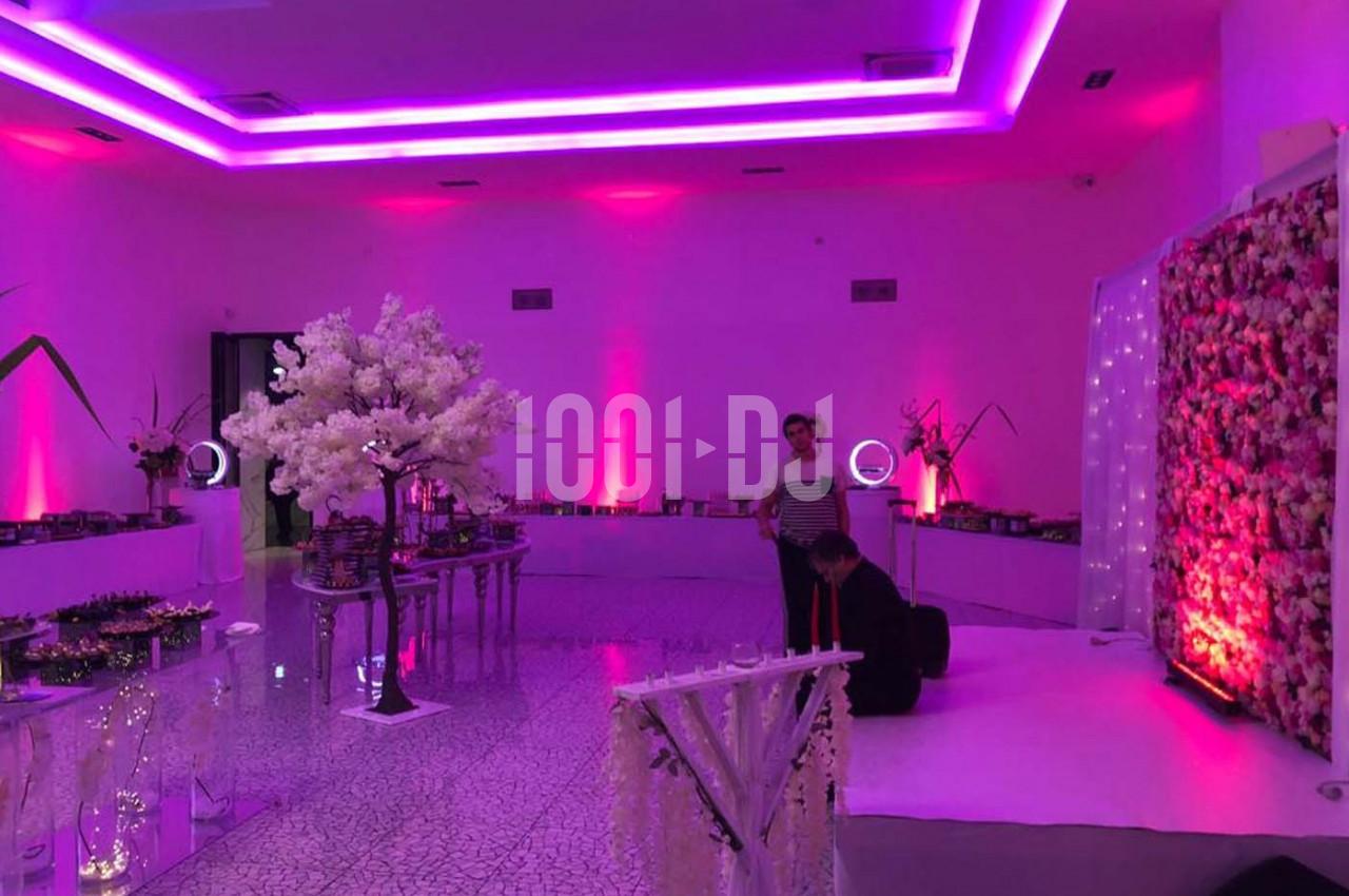 Salle décorée avec des lumières violettes, un arbre artificiel blanc et un mur floral, avec deux personnes présentes.