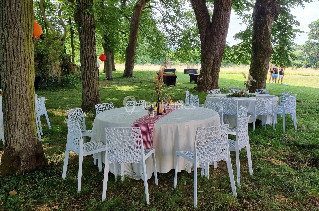 Tables rondes décorées avec nappes blanches et chaises blanches, disposées sous des arbres dans un cadre champêtre.