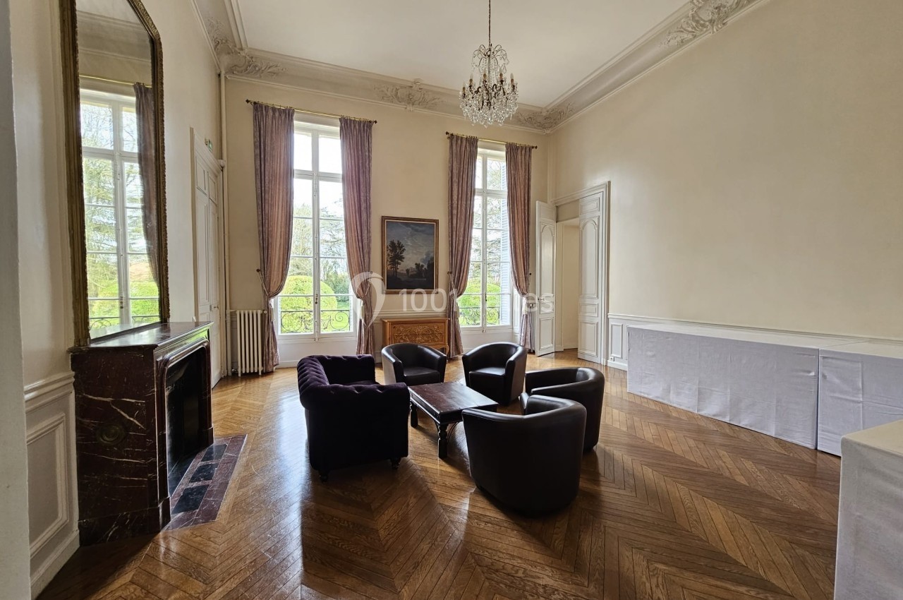 Salon lumineux avec parquet en chevrons, cheminée en marbre, fauteuils autour d'une table basse et grandes fenêtres.
