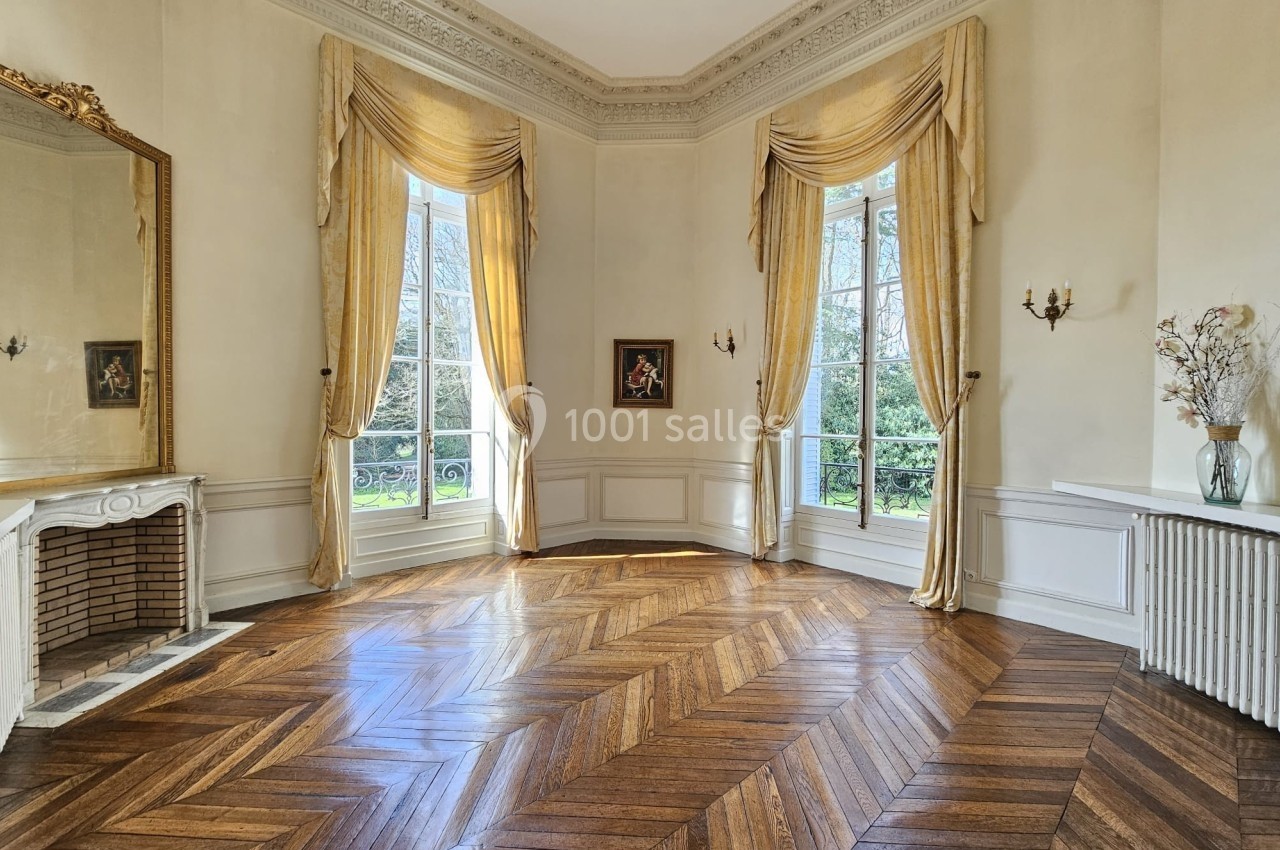 Salon lumineux avec parquet en chevrons, grandes fenêtres encadrées de rideaux dorés, cheminée et miroir décoratif.