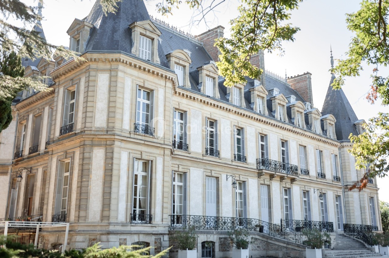 Façade d'un château en pierre claire avec des toits en ardoise, entouré de végétation et baigné de lumière naturelle.