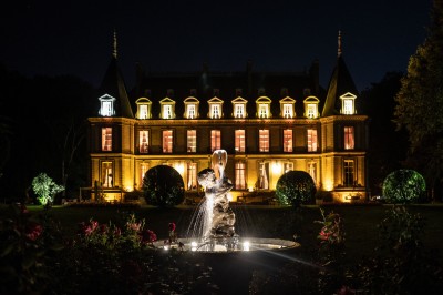 Façade d'un château illuminé la nuit avec une fontaine ornée de statues au premier plan.