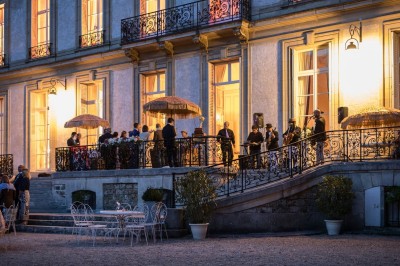 Façade d'un château illuminé la nuit avec une fontaine ornée de statues au premier plan.