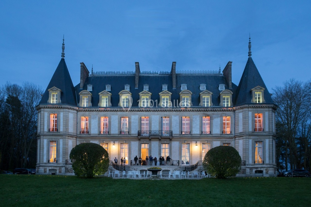 Façade éclairée d'un château au crépuscule, avec des invités rassemblés sur la terrasse et des chaises sur la pelouse.