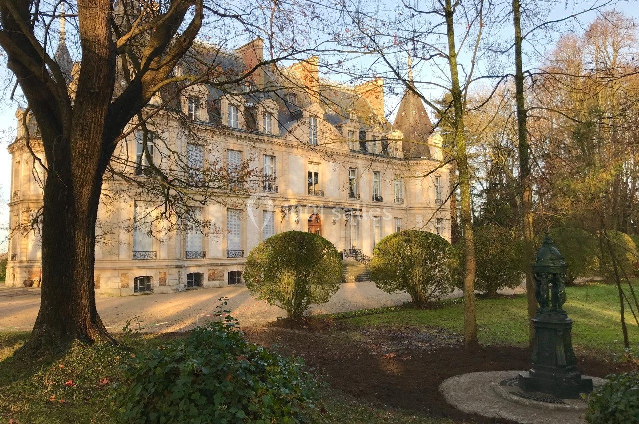 Façade d'un manoir entouré d'arbres et de buissons dans un jardin, éclairé par une lumière douce.