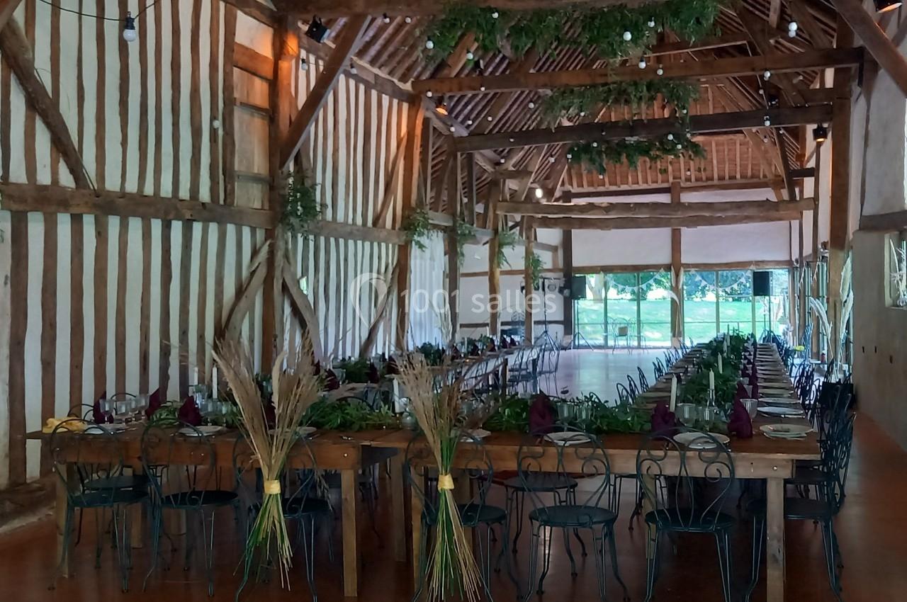 Salle de réception rustique avec poutres apparentes, tables décorées de feuillages et grandes baies vitrées donnant sur un…