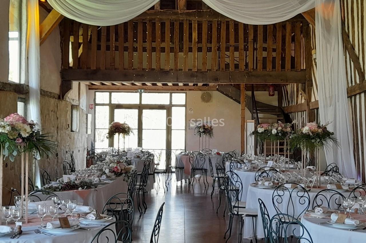 Salle de réception rustique avec tables dressées, chaises en fer forgé et décorations florales sous des drapés blancs.