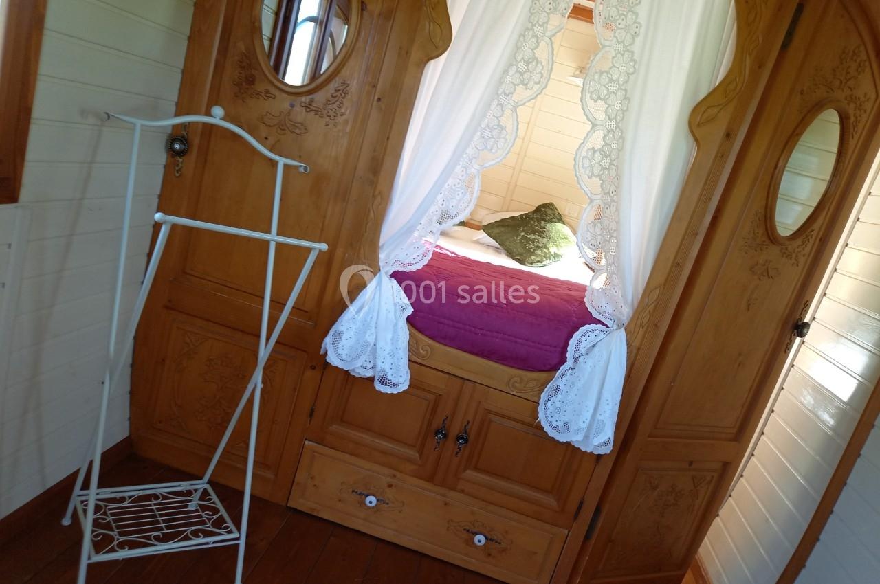 Lit avec draps blancs et coussin vert encadré par des rideaux en dentelle, dans une pièce en bois avec porte-manteau…