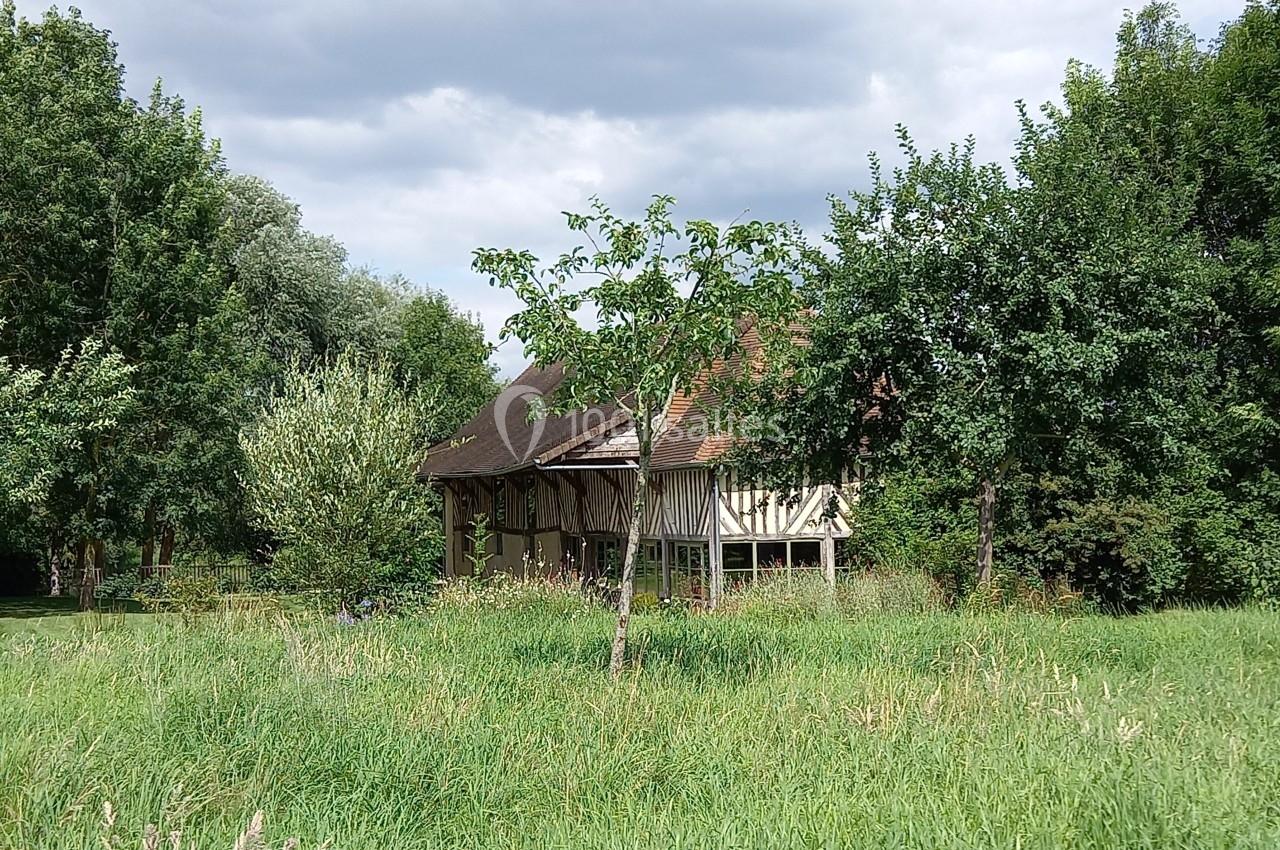 Maison à colombages entourée d'arbres et d'herbes hautes sous un ciel partiellement nuageux.