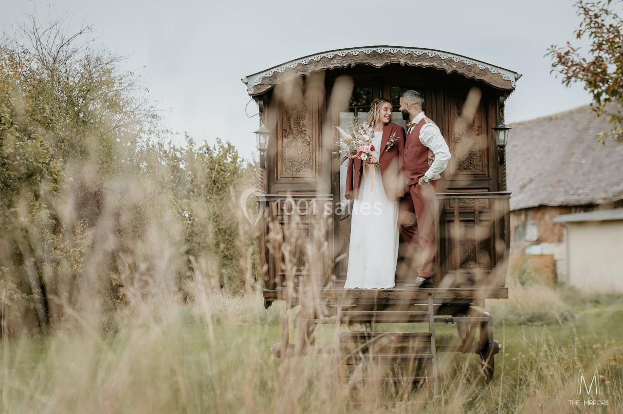 Un couple en tenue de mariage pose sur la plateforme d'une roulotte en bois dans un cadre champêtre.