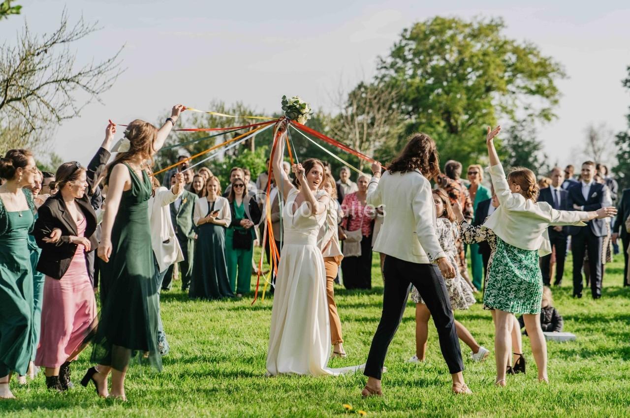 Des personnes dansent en cercle autour d'une mariée tenant des rubans colorés, dans un jardin ensoleillé.