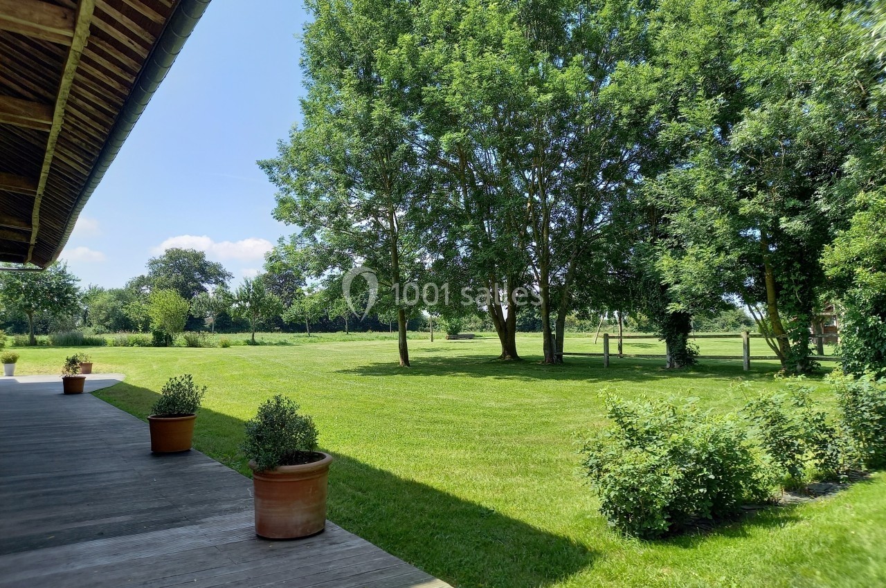 Terrasse en bois bordée de pots de plantes, donnant sur un grand jardin verdoyant avec des arbres sous un ciel bleu.