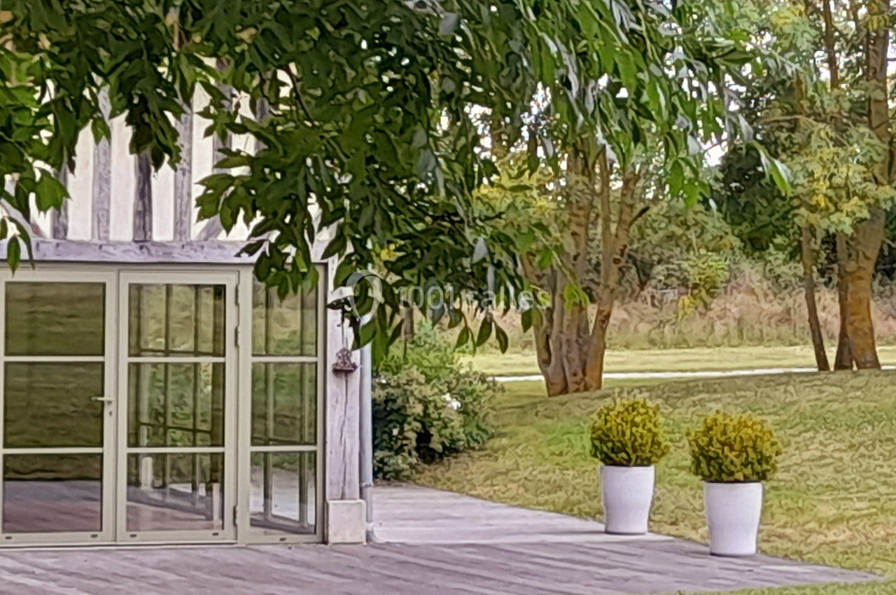 Façade vitrée d'une maison en bois avec terrasse en bois, deux pots de buis et un jardin arboré en arrière-plan.