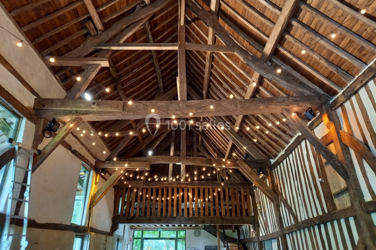 Charpente en bois apparente d'une grange rénovée, éclairée par des guirlandes lumineuses suspendues.