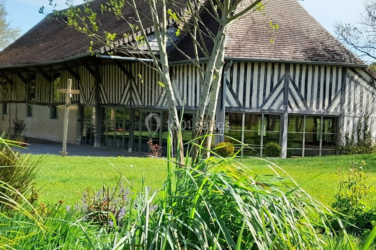 Bâtiment à colombages avec toit en pente, entouré de verdure et d'arbres, sous un ciel clair.
