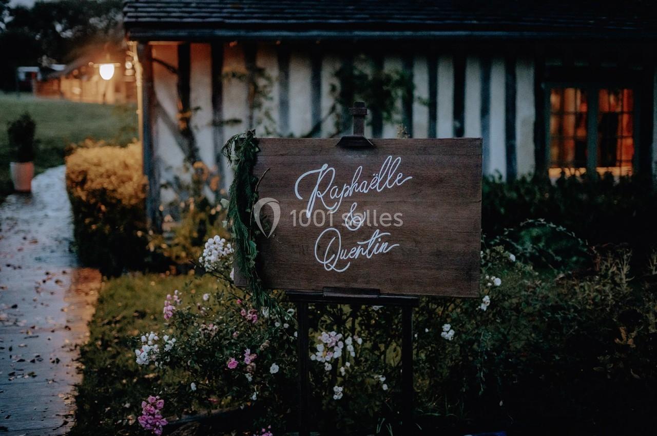 Panneau en bois décoré de fleurs, portant les prénoms ’Raphaëlle & Quentin’, devant une maison à colombages.