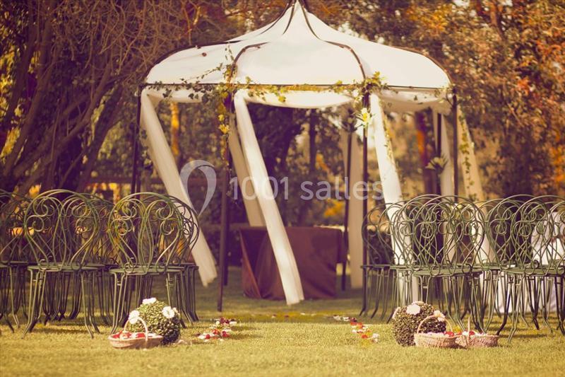 Tente blanche décorée dans un jardin, entourée de chaises en fer forgé et de pétales de fleurs au sol.