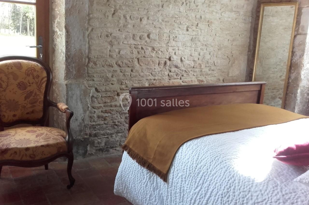 Chambre avec mur en pierre, lit recouvert d'un plaid jaune, fauteuil ancien et grand miroir encadré.