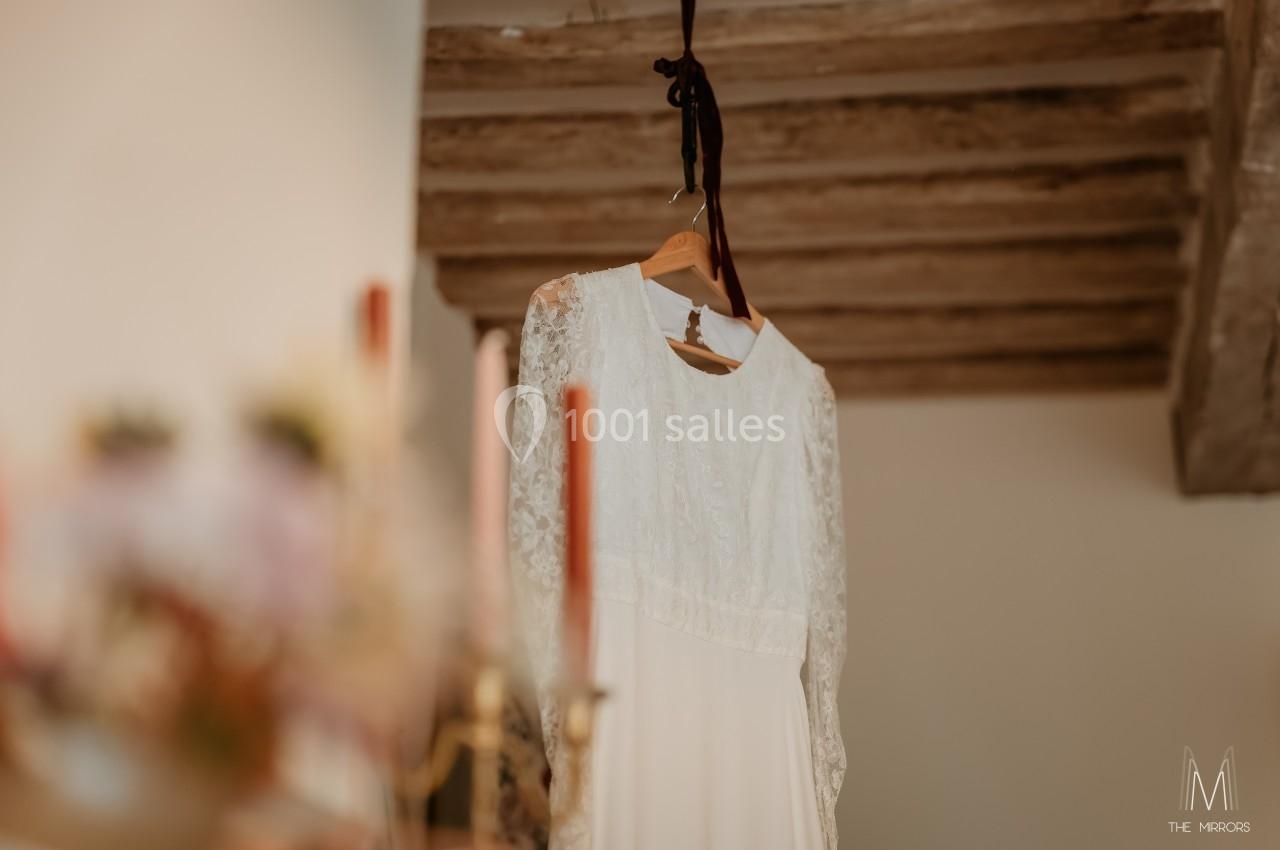 Robe de mariée blanche en dentelle suspendue sur un cintre, avec des poutres en bois en arrière-plan.