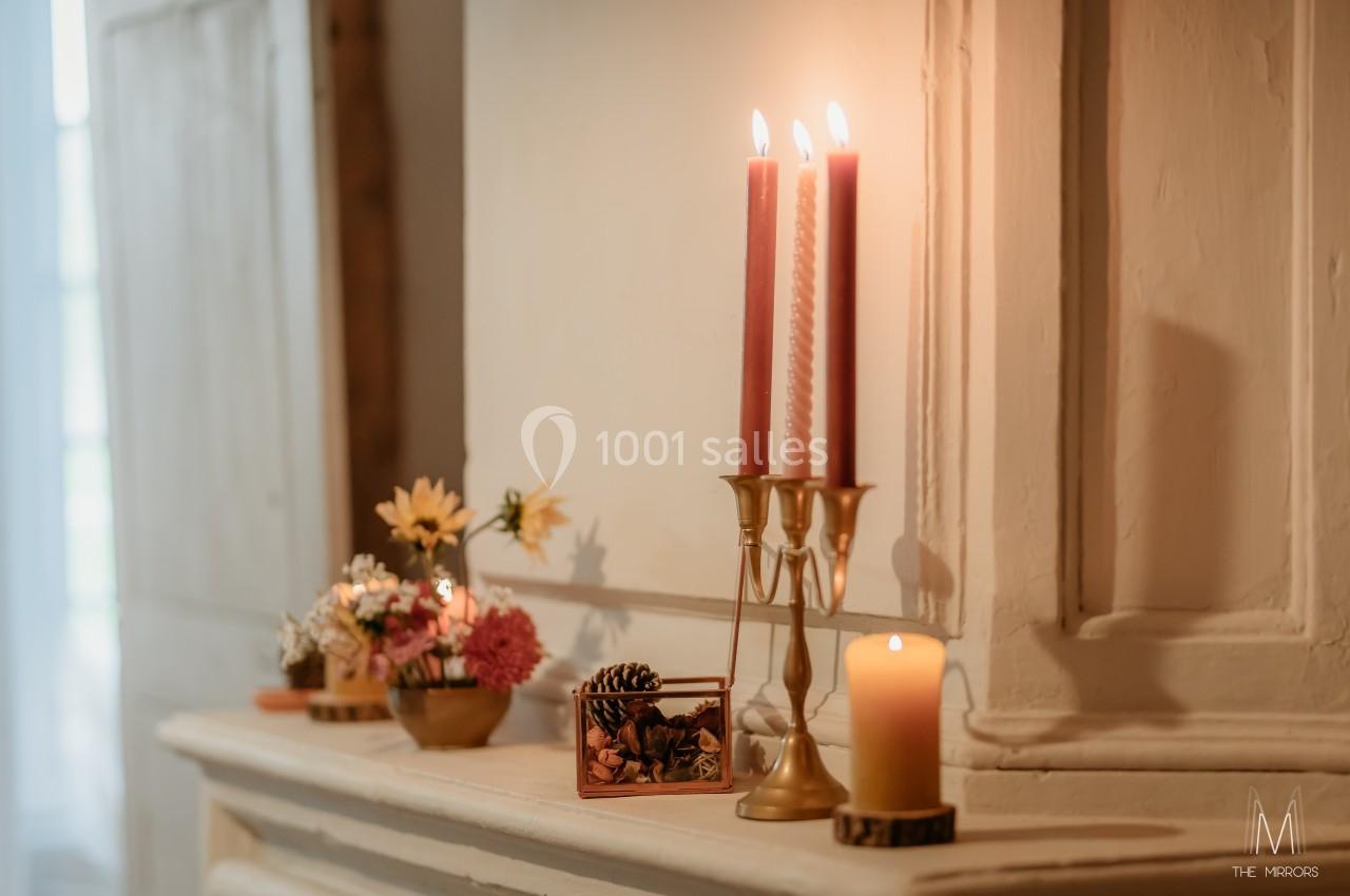 Bougeoir doré avec trois bougies allumées, fleurs et décorations posées sur une cheminée en bois clair.