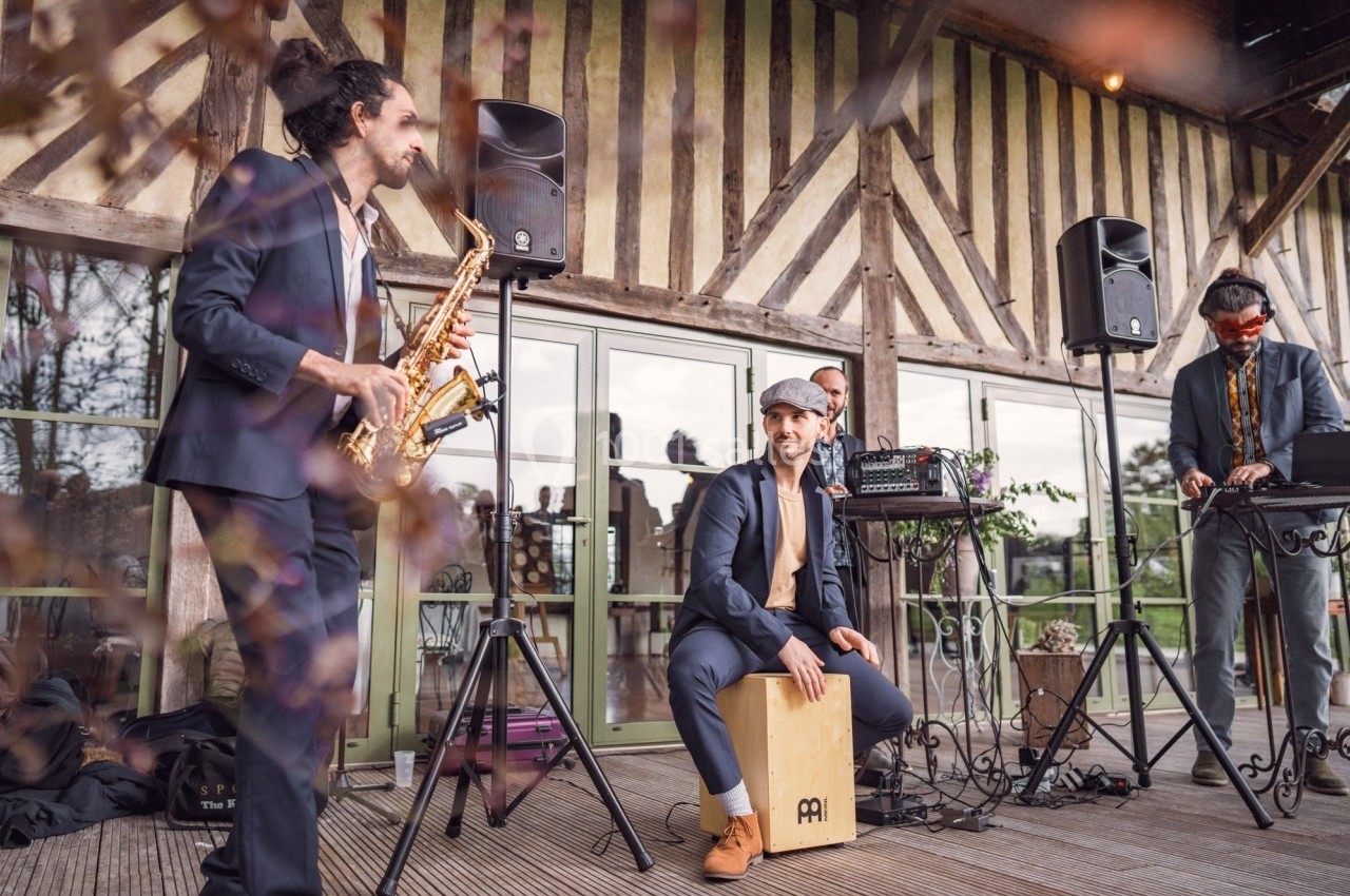 Un groupe de musiciens joue en extérieur sous une terrasse couverte, avec saxophone, cajón et matériel audio.