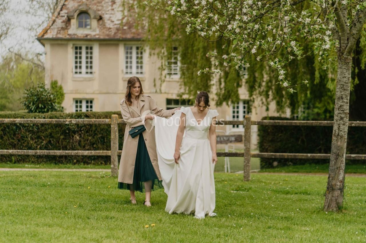 Une femme aide une mariée à porter sa robe dans un jardin verdoyant devant une maison ancienne.