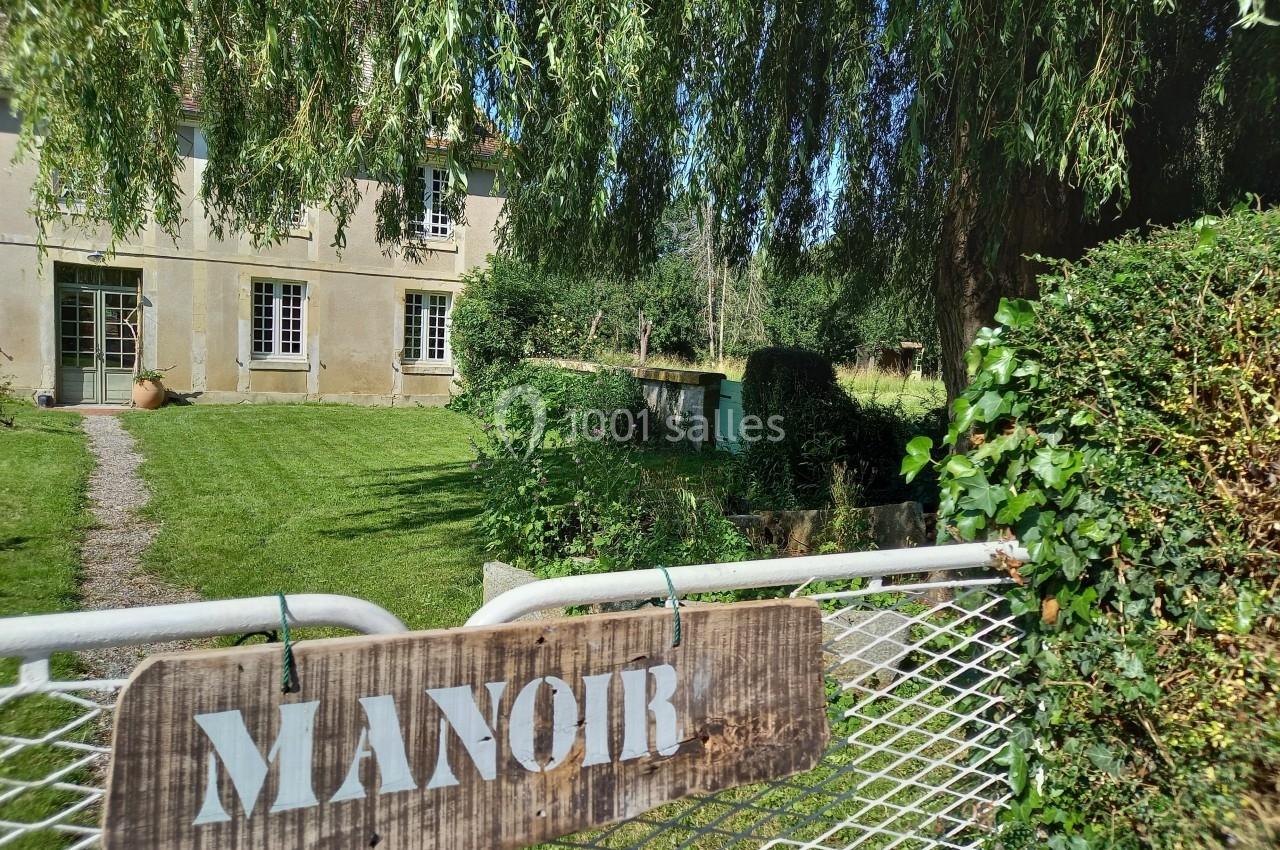 Vue d'un manoir en pierre entouré de verdure, avec une enseigne en bois fixée sur un portail métallique.