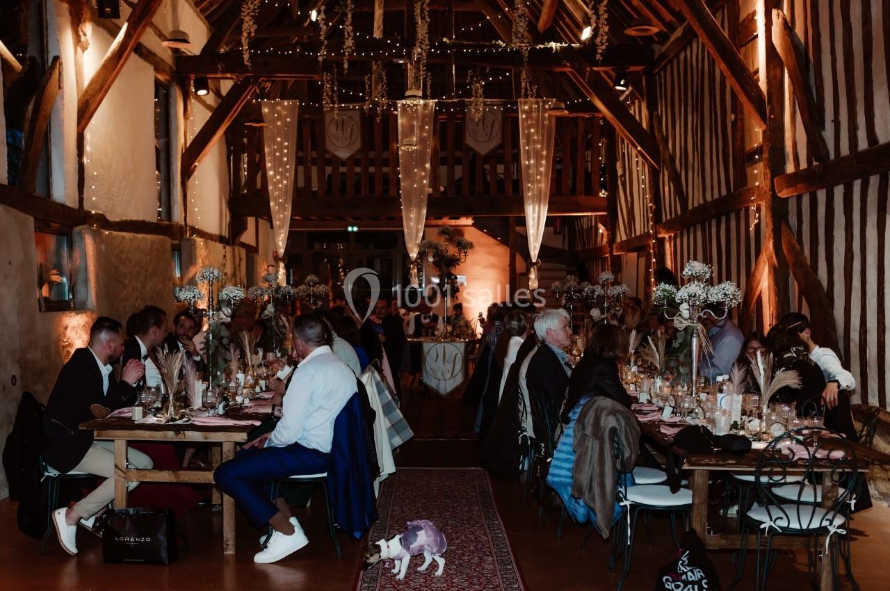 Salle de réception rustique avec des invités attablés, décorée de guirlandes lumineuses et de fleurs suspendues.
