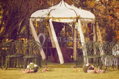 Salle de réception rustique avec tables rondes dressées, chaises en fer forgé et drapés blancs suspendus au plafond.