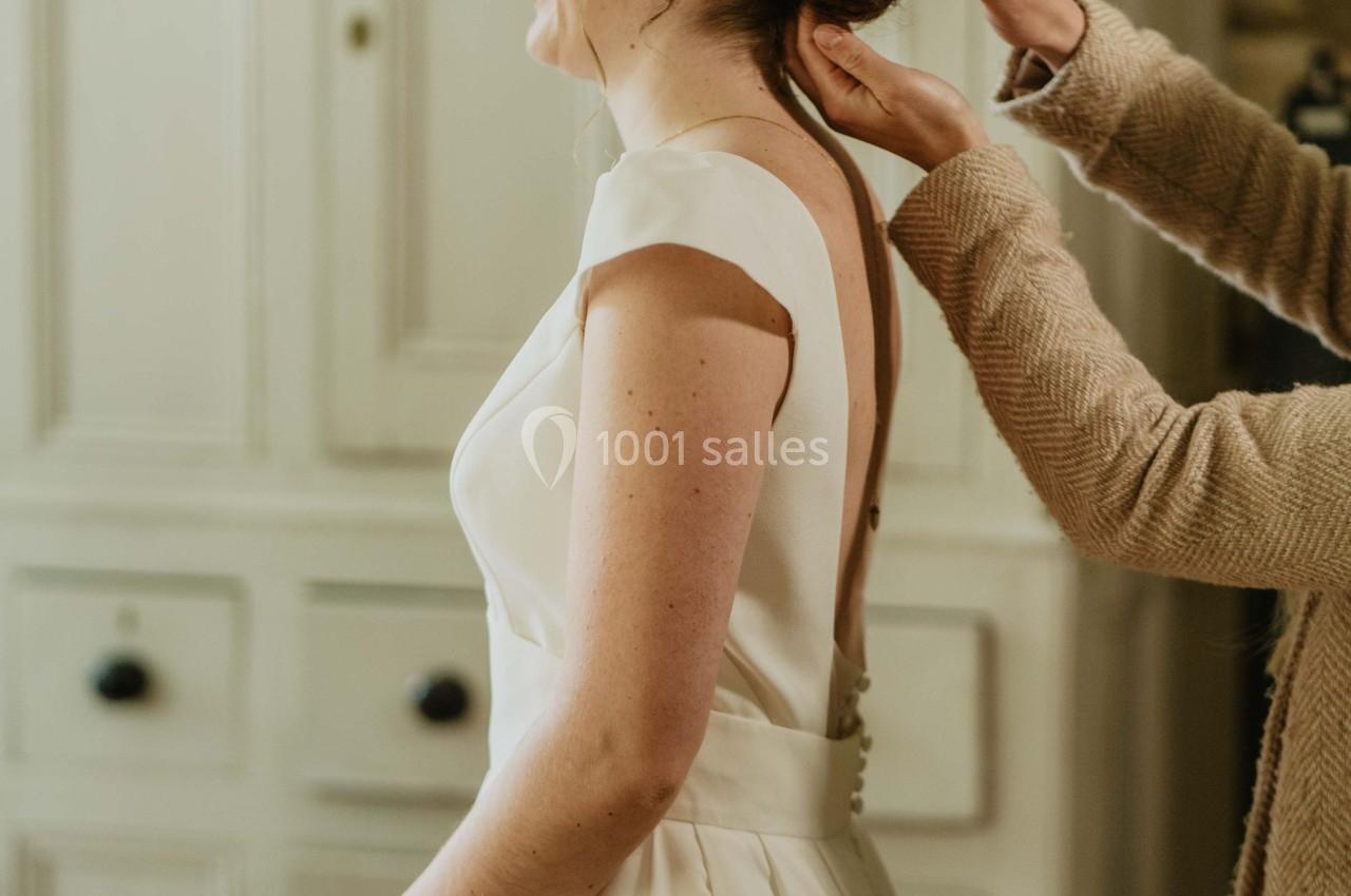 Une femme en robe de mariée blanche se fait ajuster la coiffure par une autre personne dans une pièce lumineuse.