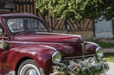 Voiture ancienne rouge décorée de fleurs blanches et feuillages, stationnée devant un bâtiment à colombages.