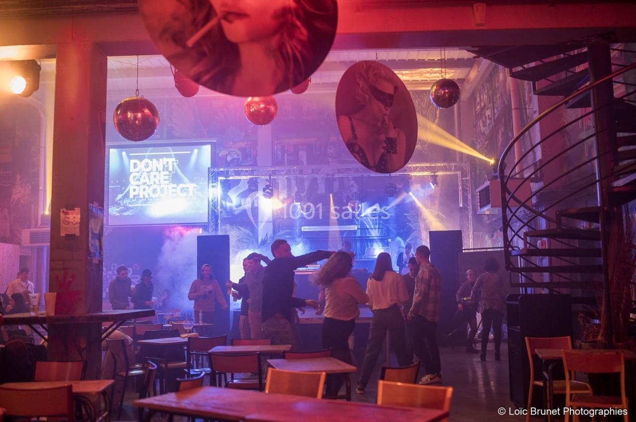 Des personnes dansent dans une salle éclairée par des lumières colorées, avec des tables et des décorations suspendues.